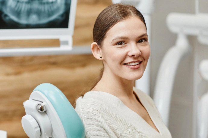 Woman smiling in dental office chair with overhead light and x-ray visible in background.