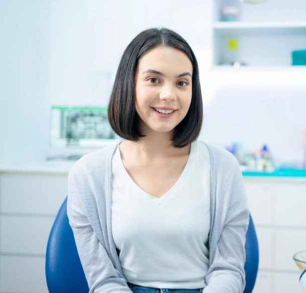 Smiling woman sitting in dental office chair with shelves of supplies visible in background.