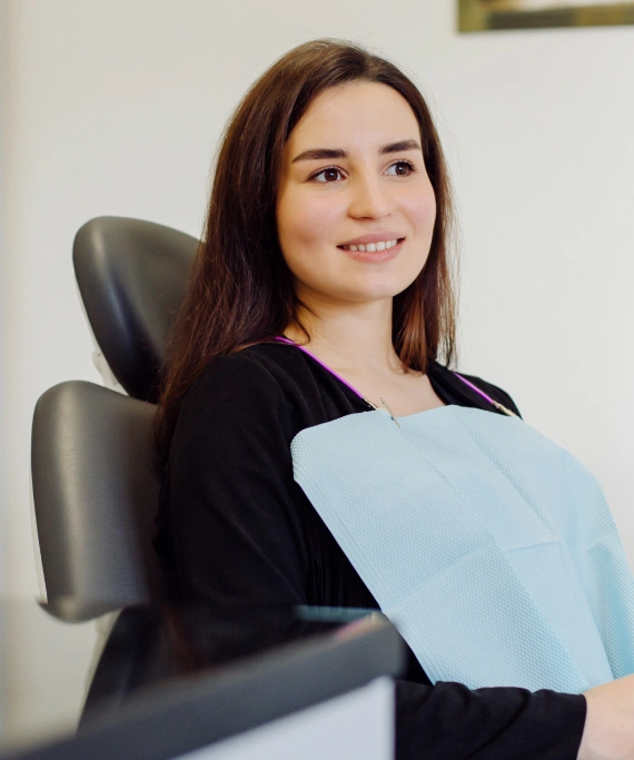 Young woman smiling at dental office, holding light blue bib, seated in dental chair.