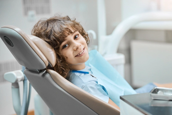 Young child seated in dental chair wearing blue bib in modern dental office setting.