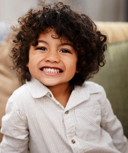 Young child with curly hair smiling brightly wearing light striped shirt indoors.