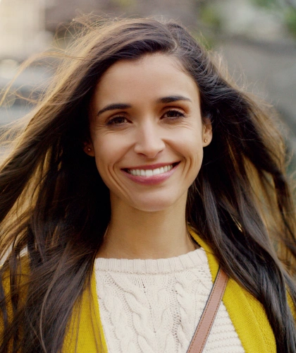 Young woman smiling at camera outdoors wearing yellow cardigan and white shirt.
