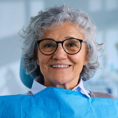 Smiling senior woman wearing glasses and blue jacket in dental office setting.