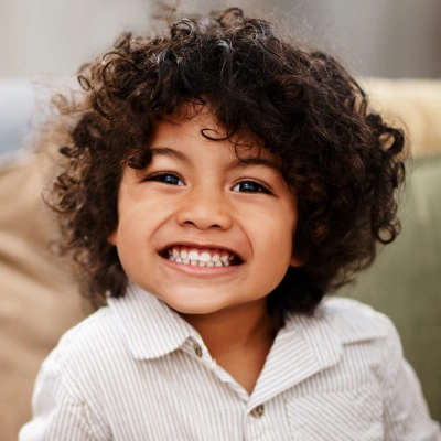 Young child with curly hair smiling brightly in white collared shirt, indoor setting.