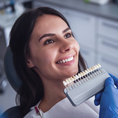 Smiling dental professional in blue gloves holding tooth shade guide in modern dental office.