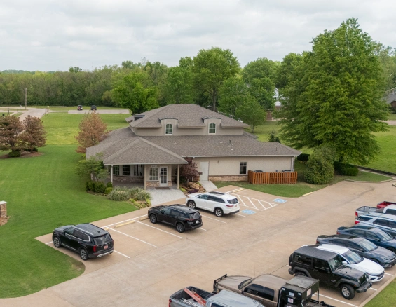 Modern office building exterior with parking lot, landscaped lawn, and surrounding trees on an overcast day.